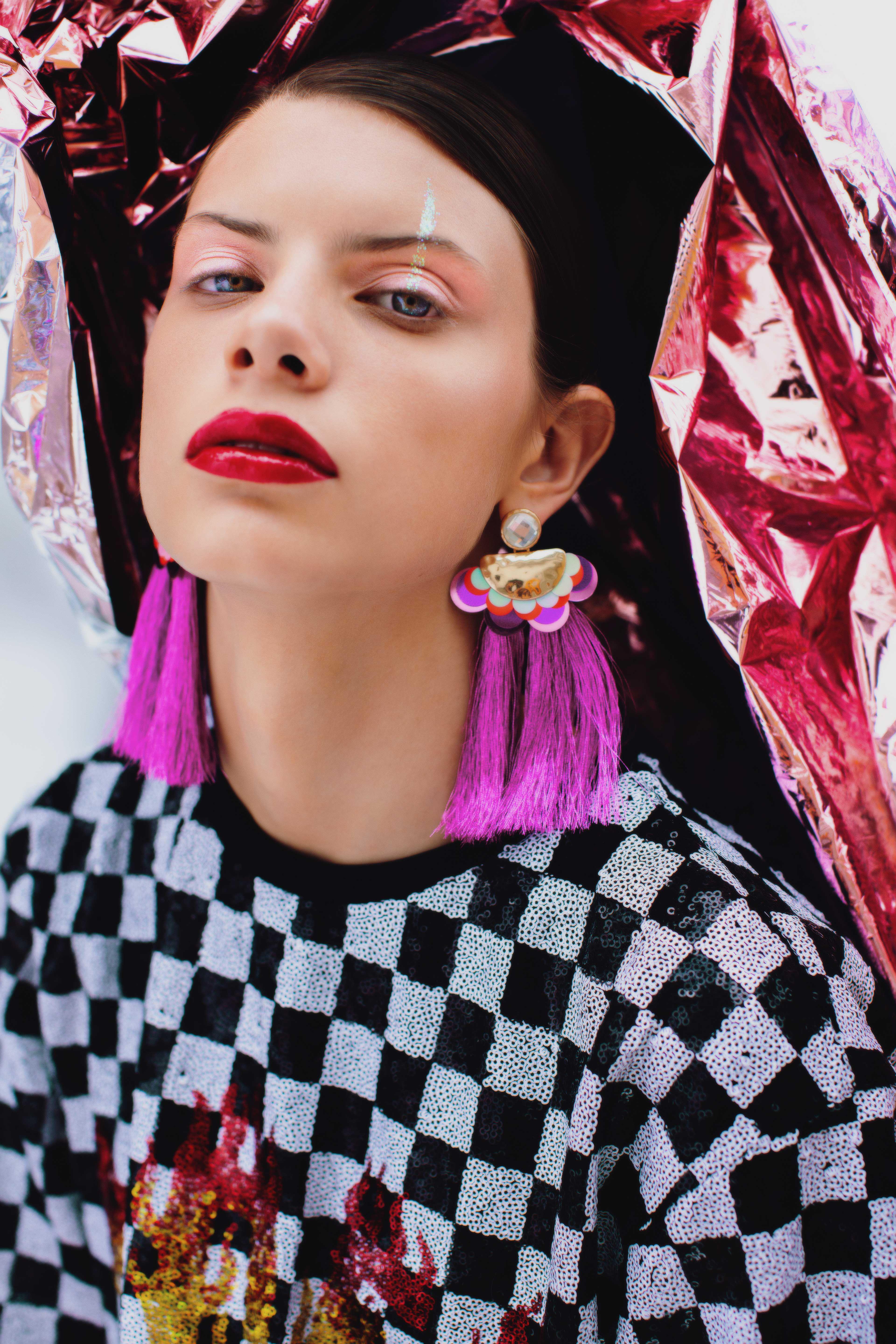 portrait of a woman with a plaid tshirt and purple earrings and a strip of glitter above her eyebrow.