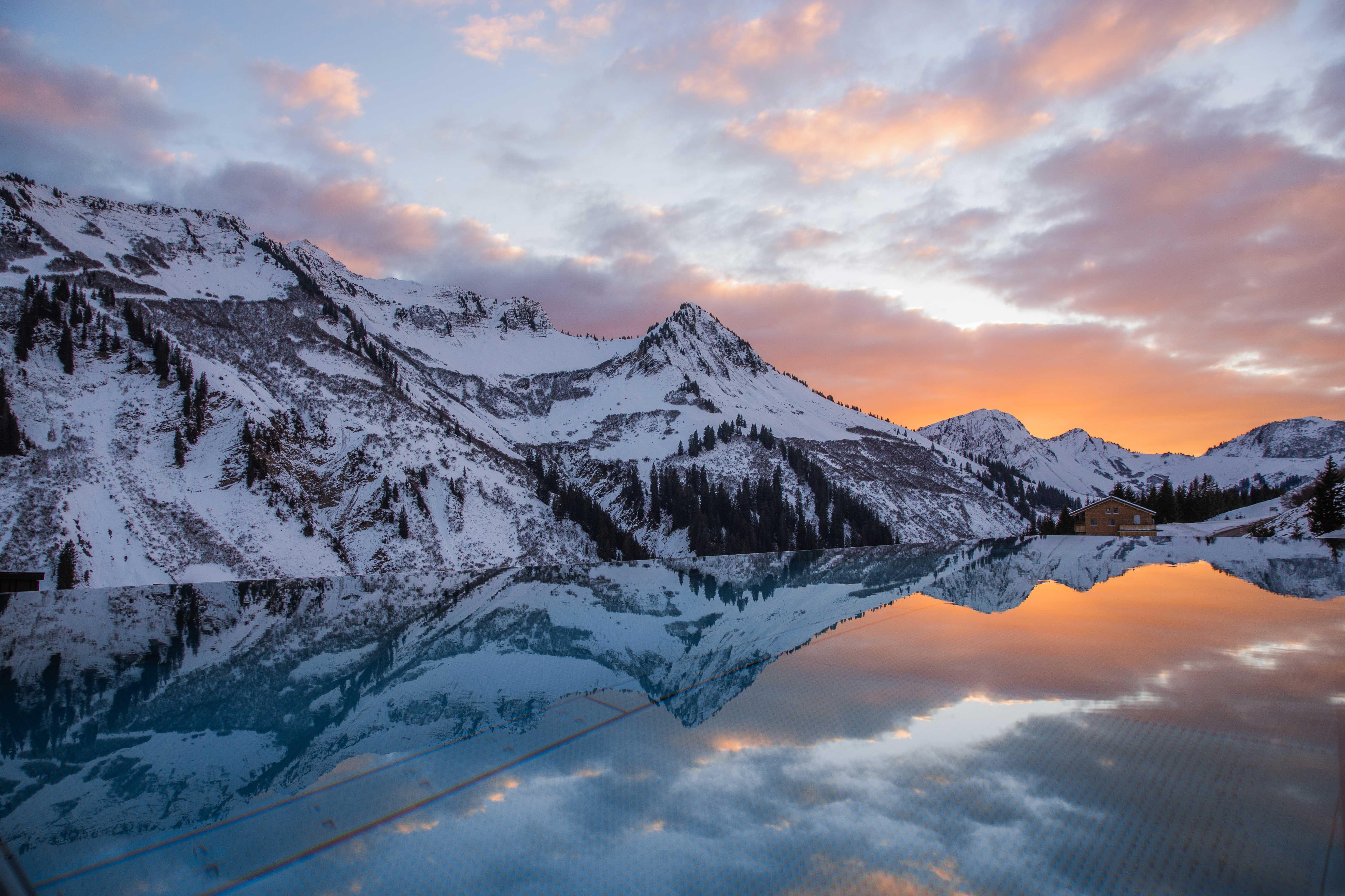 photo of a snowy mountain range in the sunset which is reflected in the calm water of the outdoor infinity pool.