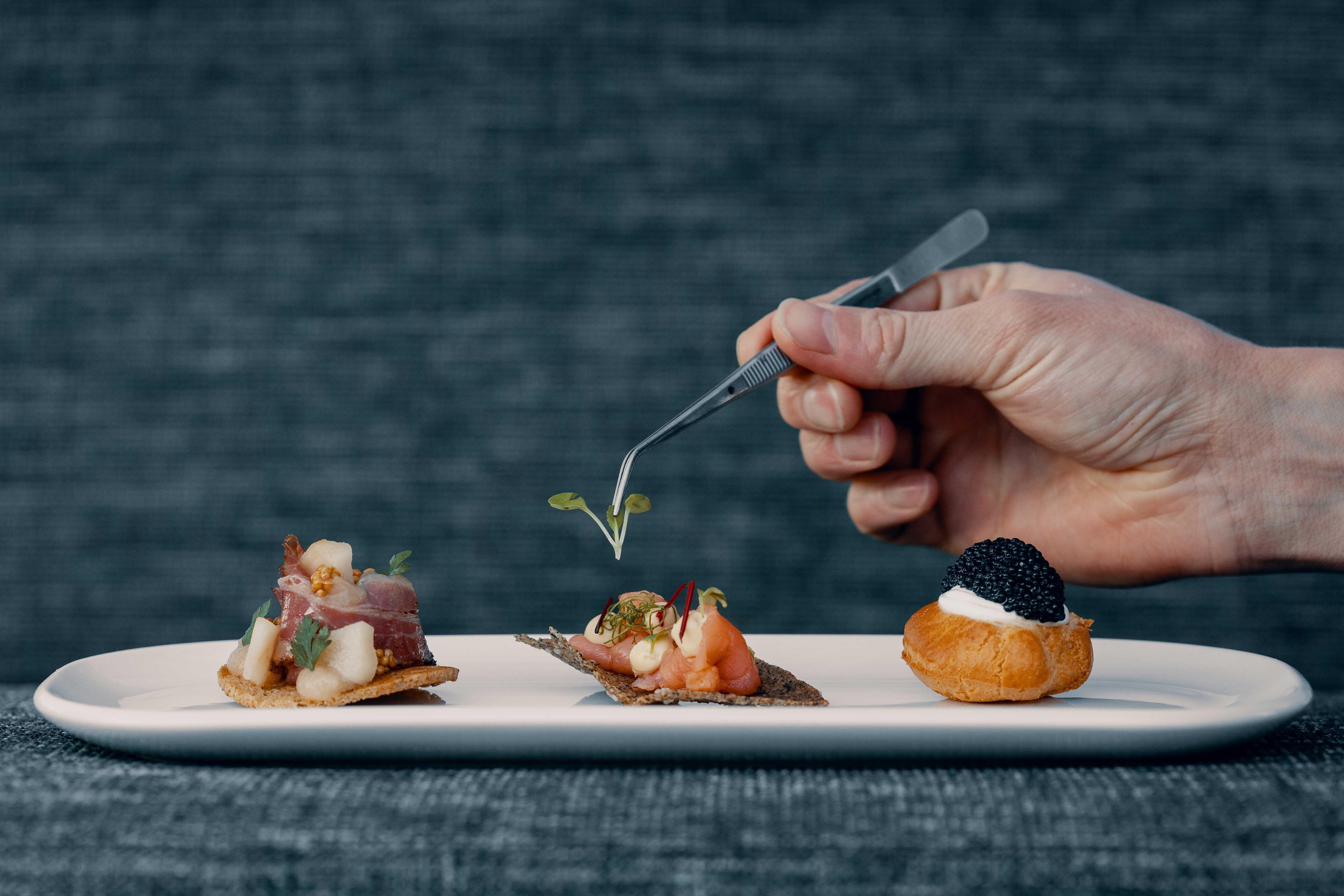 close-up of a hand using tweezers to place a small leaf on an appetizer arranged on a plate.