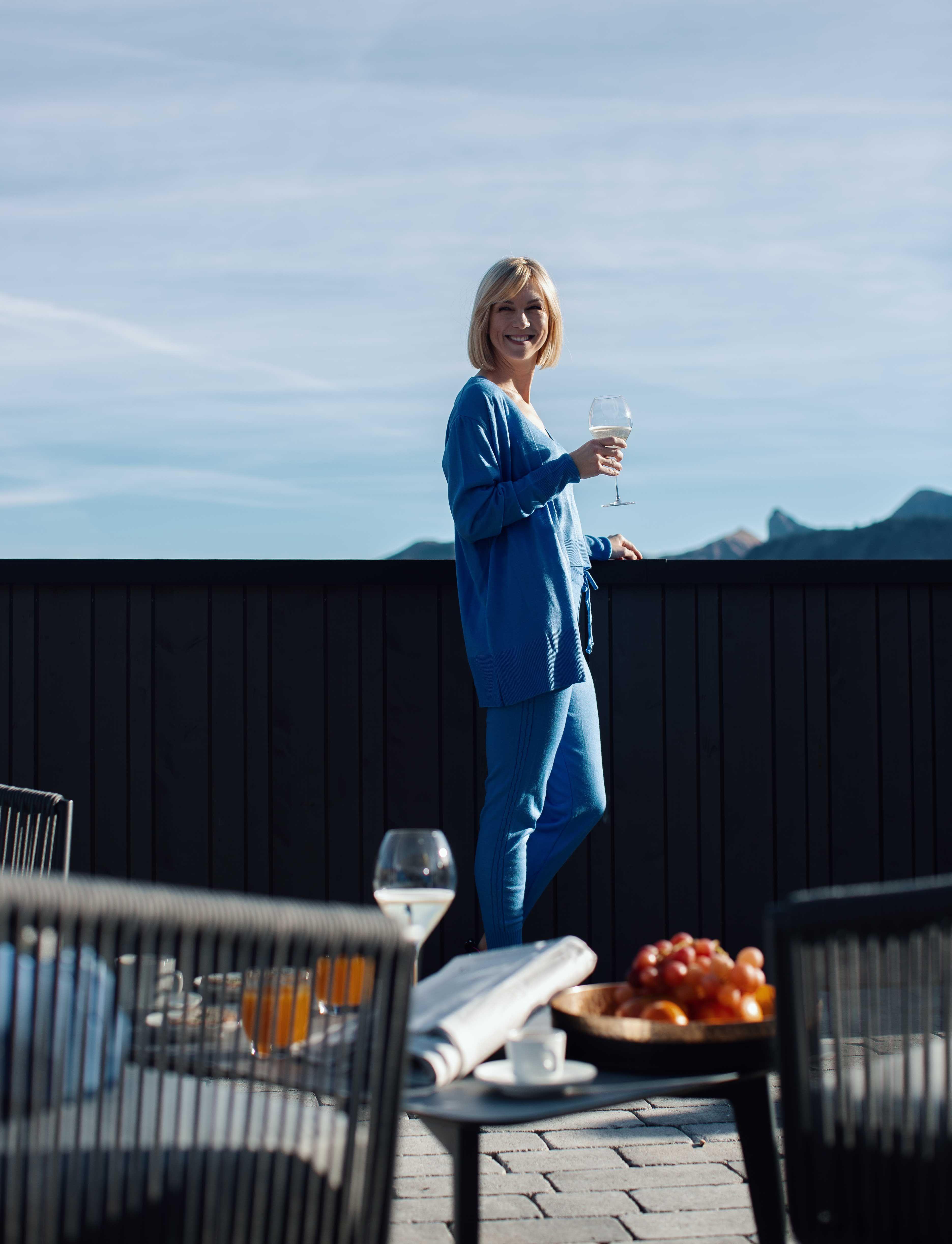 a blonde woman stands on a roof terrace, in the foreground you can see a small breakfast table with fruit, coffee and newspaper. the woman stands on the railing, smiles at us and holds a prosecco glass in her hand.