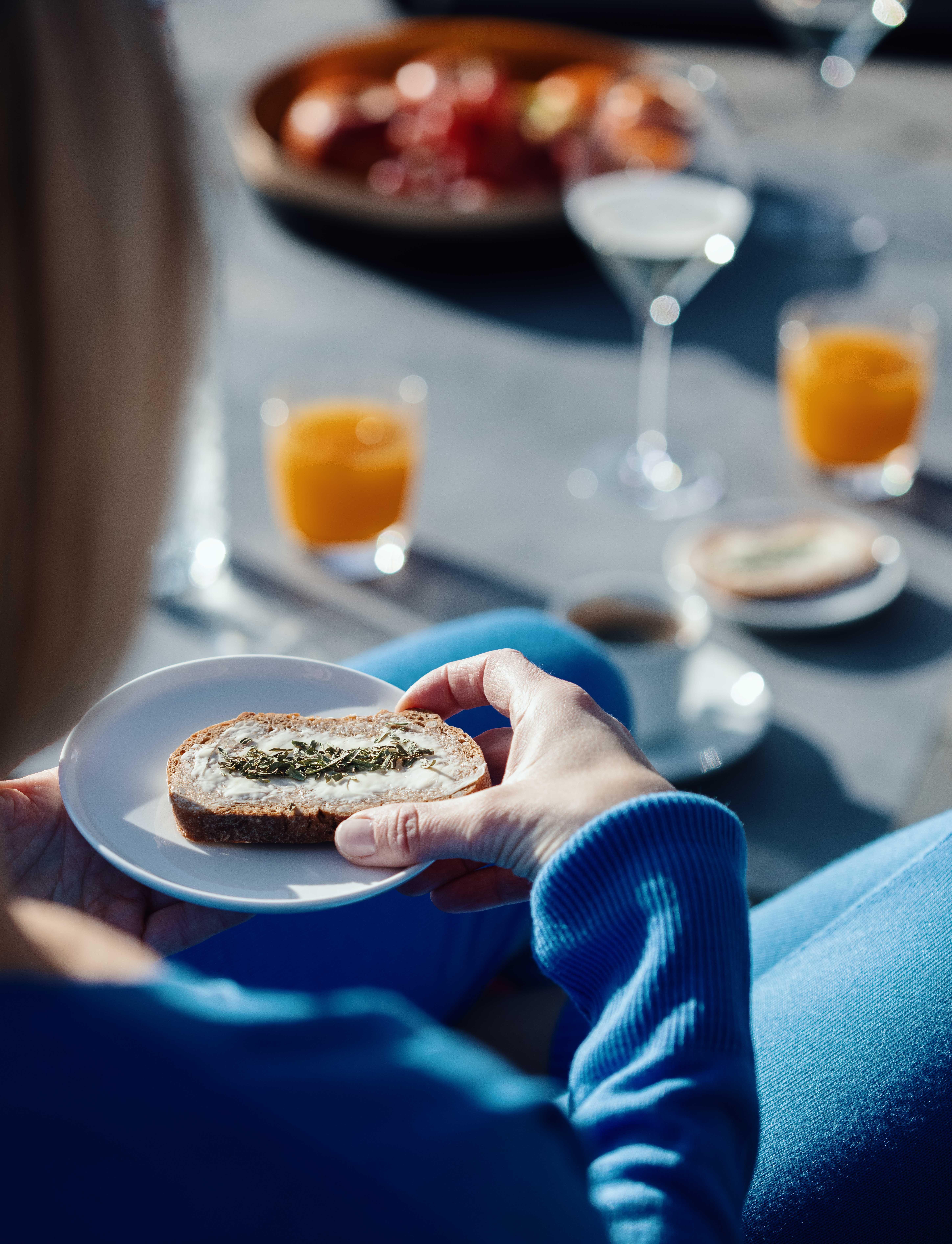 view over the shoulder of a woman holding her breakfast bread, in the background we see the breakfast table with fruit, coffee and orange juice.