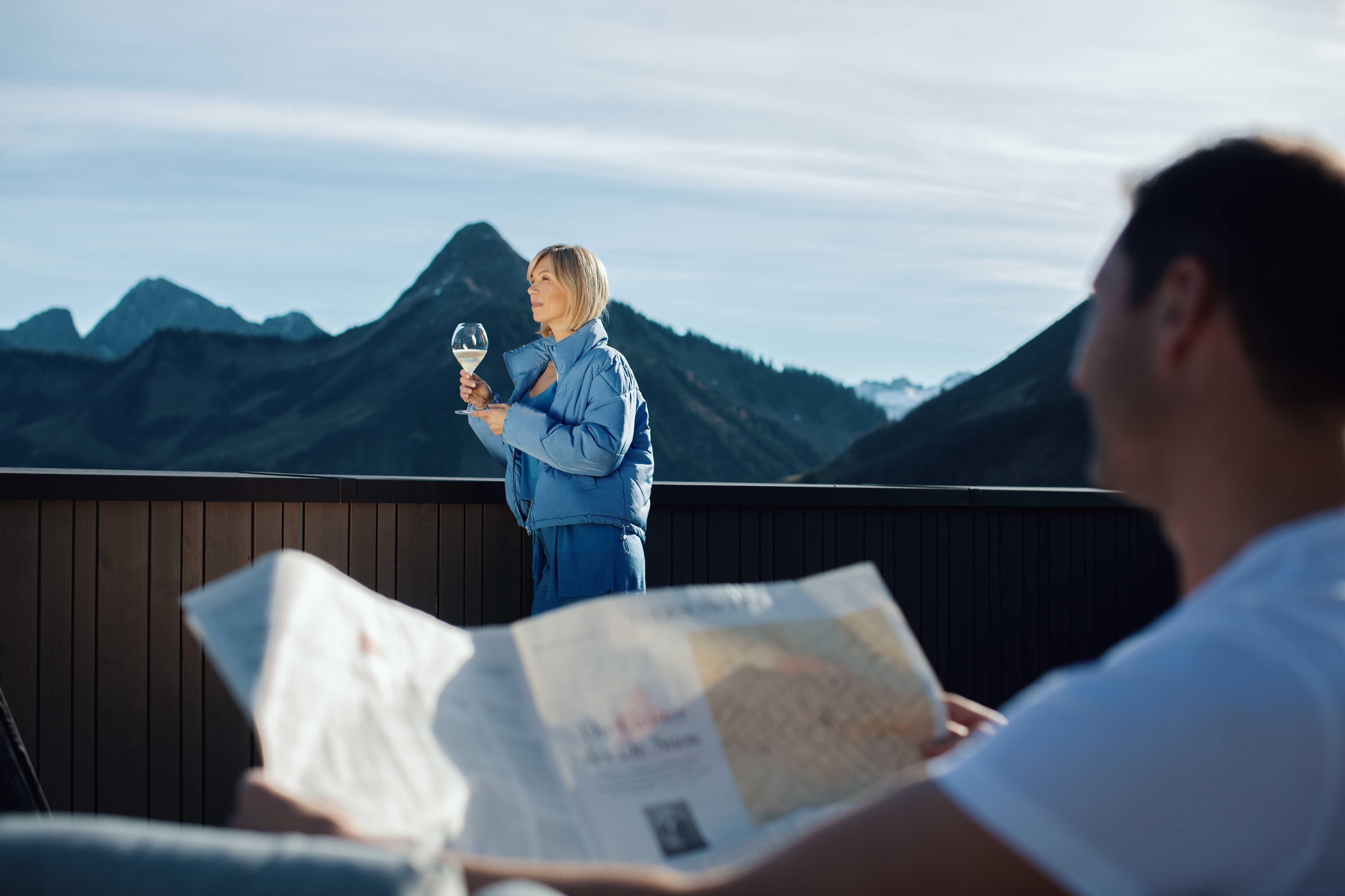 a woman stands on a roof terrace with a view of the mountains and holds a glass of prosecco in her hand. a man sits in the foreground and reads the newspaper.