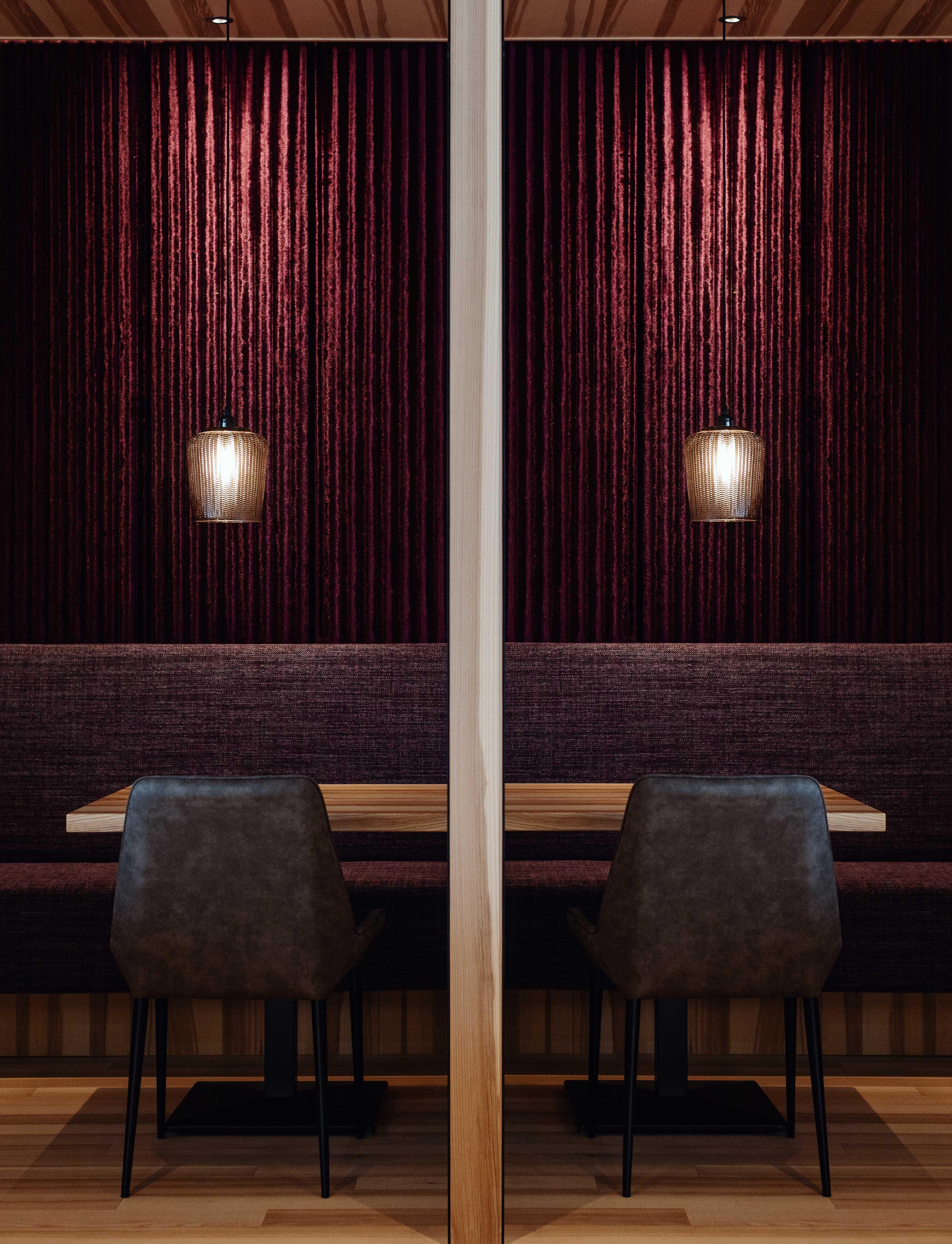 interior design shot of a restaurant, you see a table with two chairs and two lamps hanging over it in front of a velvet covered purple wall.