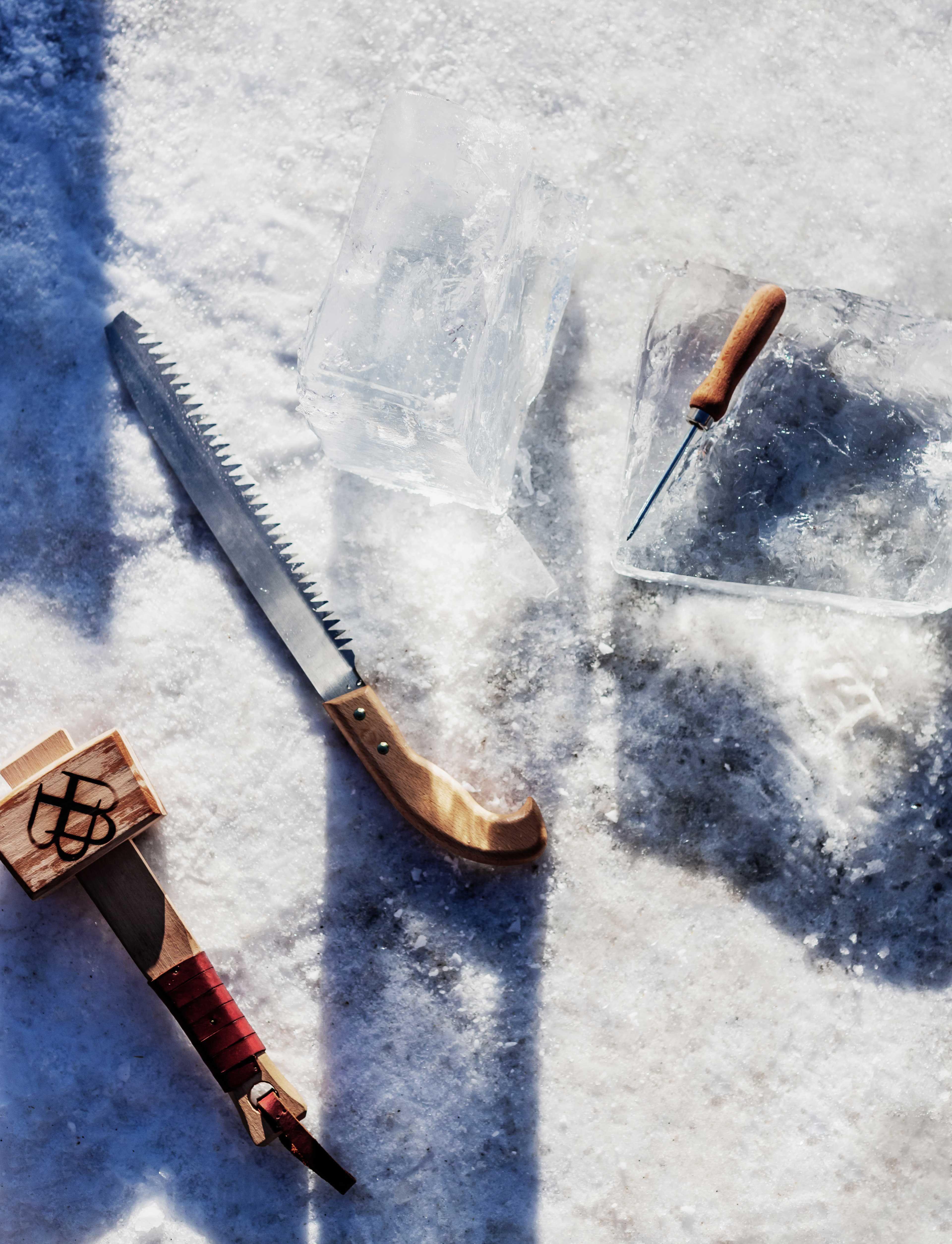 two large blocks of ice lie on the ground on snow, next to them various tools such as an ice saw and a hammer. 