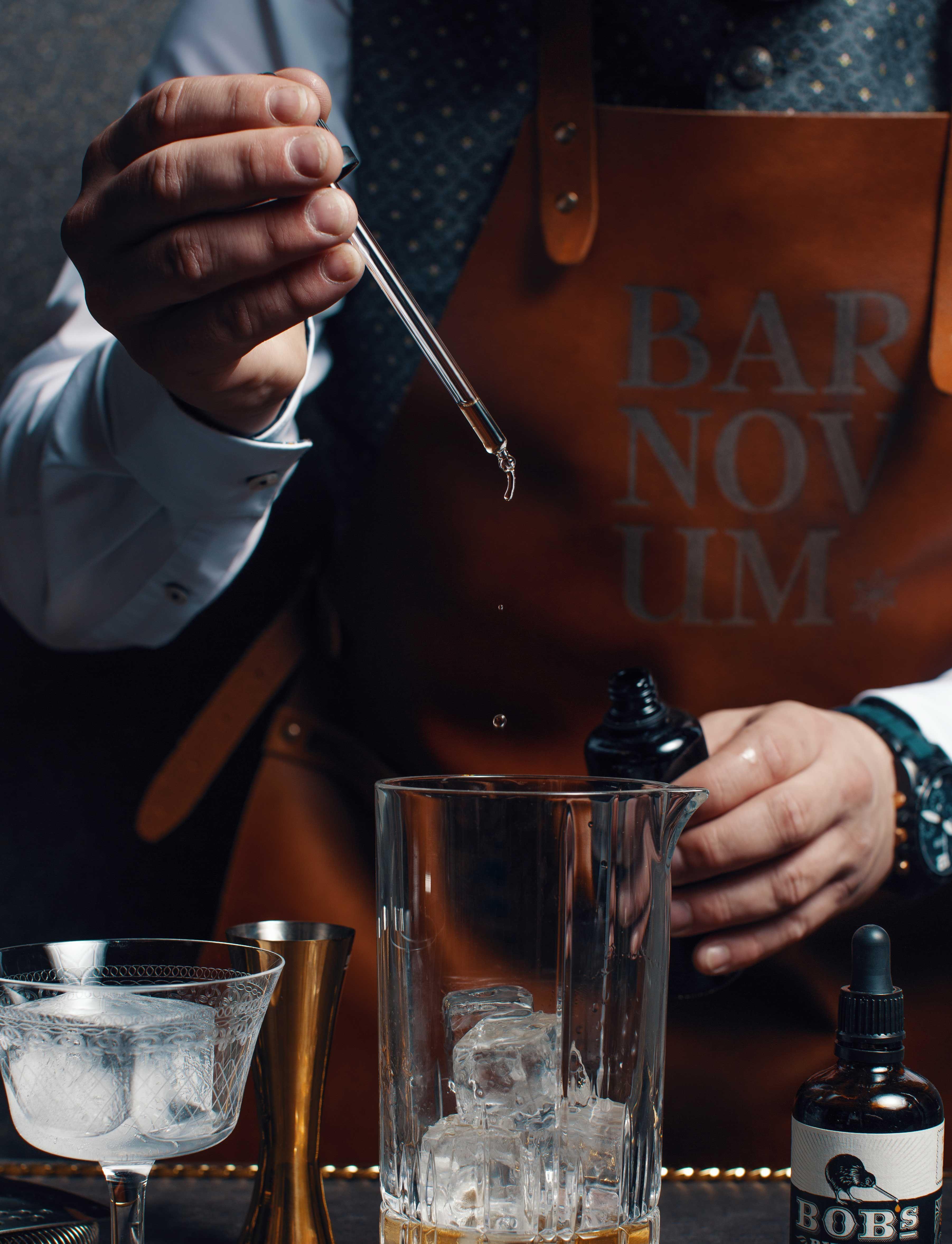 closeup of a bartender with leather apron who puts some drops of liquid into a glass with a pipette.