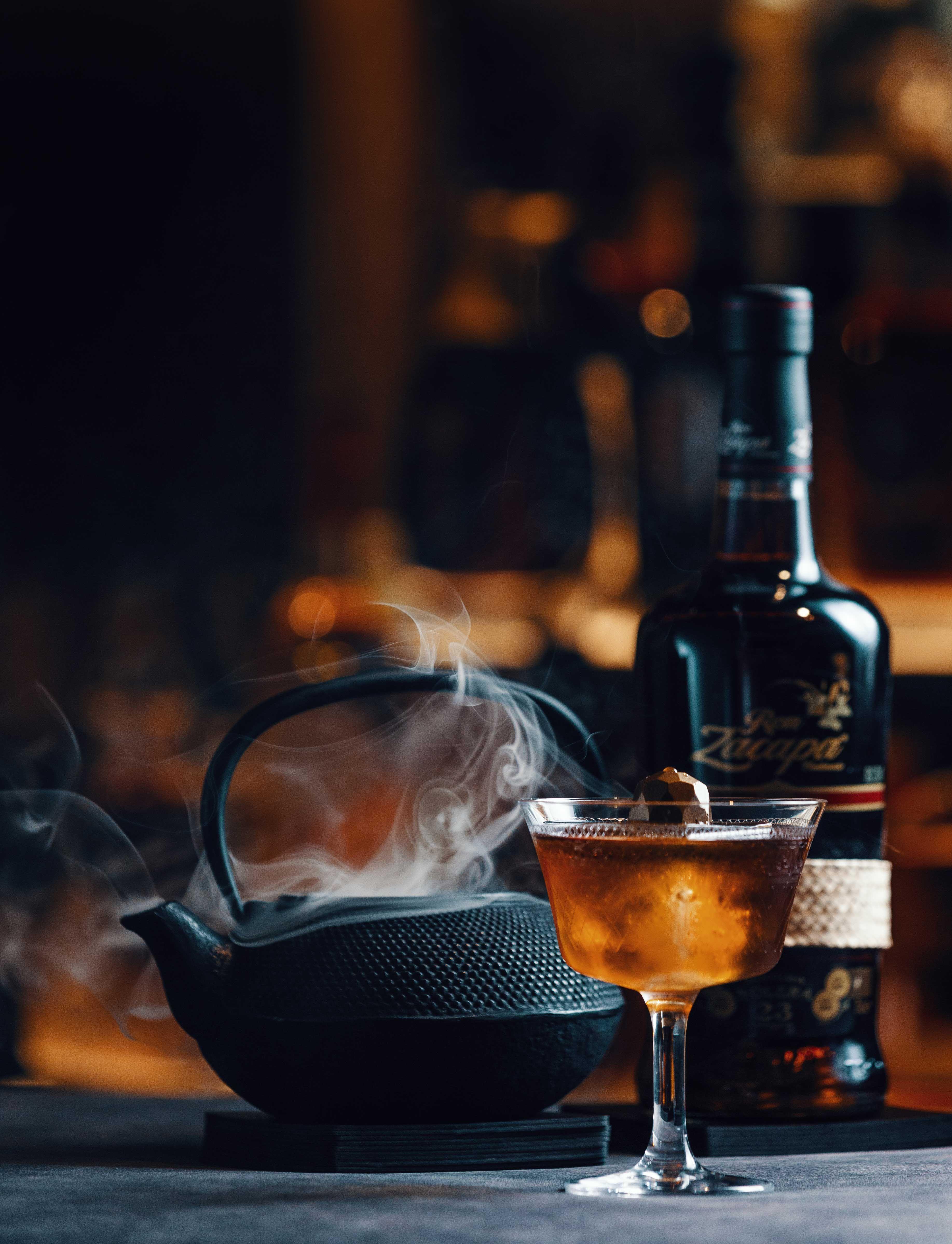 a filled cocktail glass stands in front of a heavily smoking teapot, next to it is a bottle of ron zacapa.