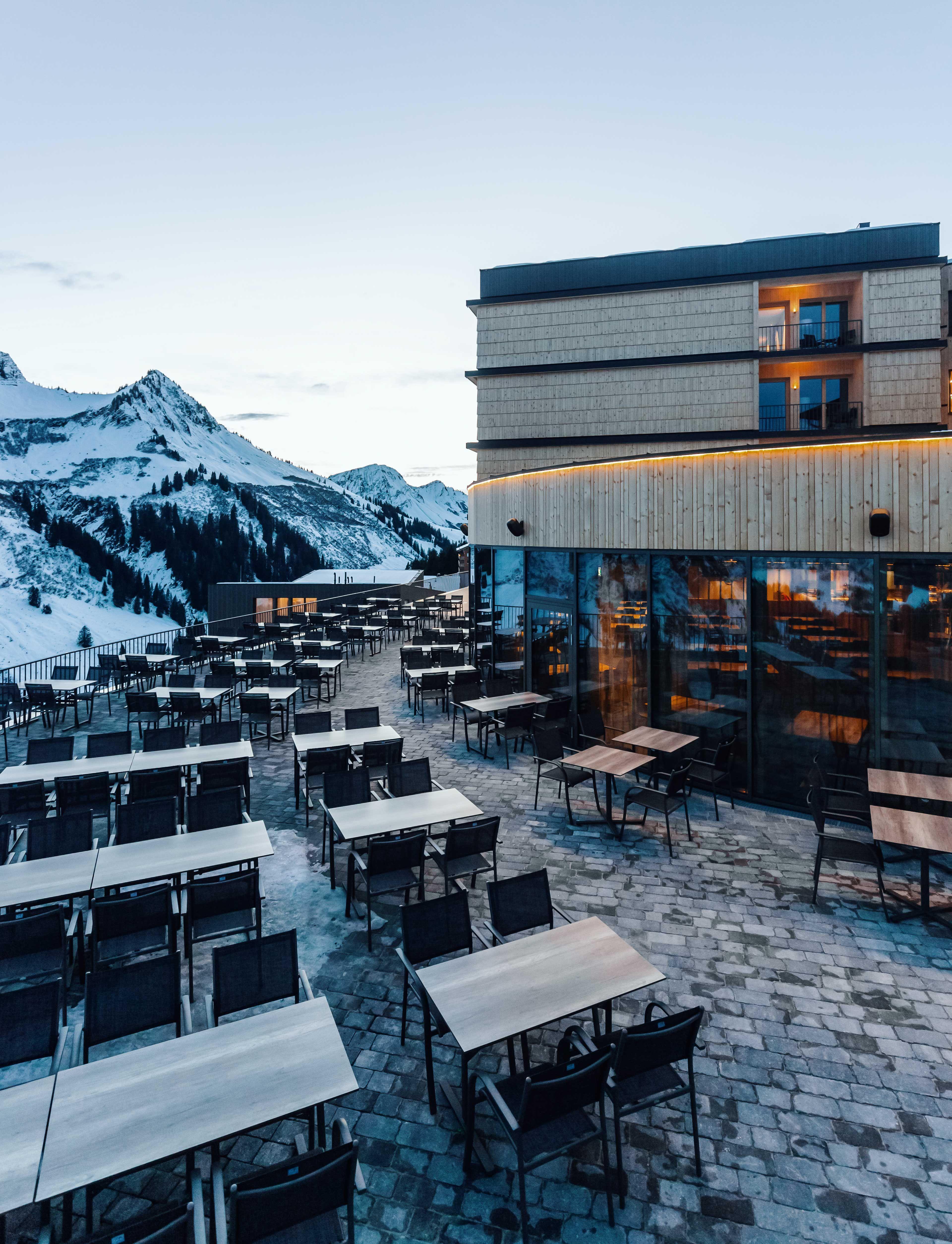 Exterior shot of a hotel in the middle of snowy mountains with a terrace arranged with tables.