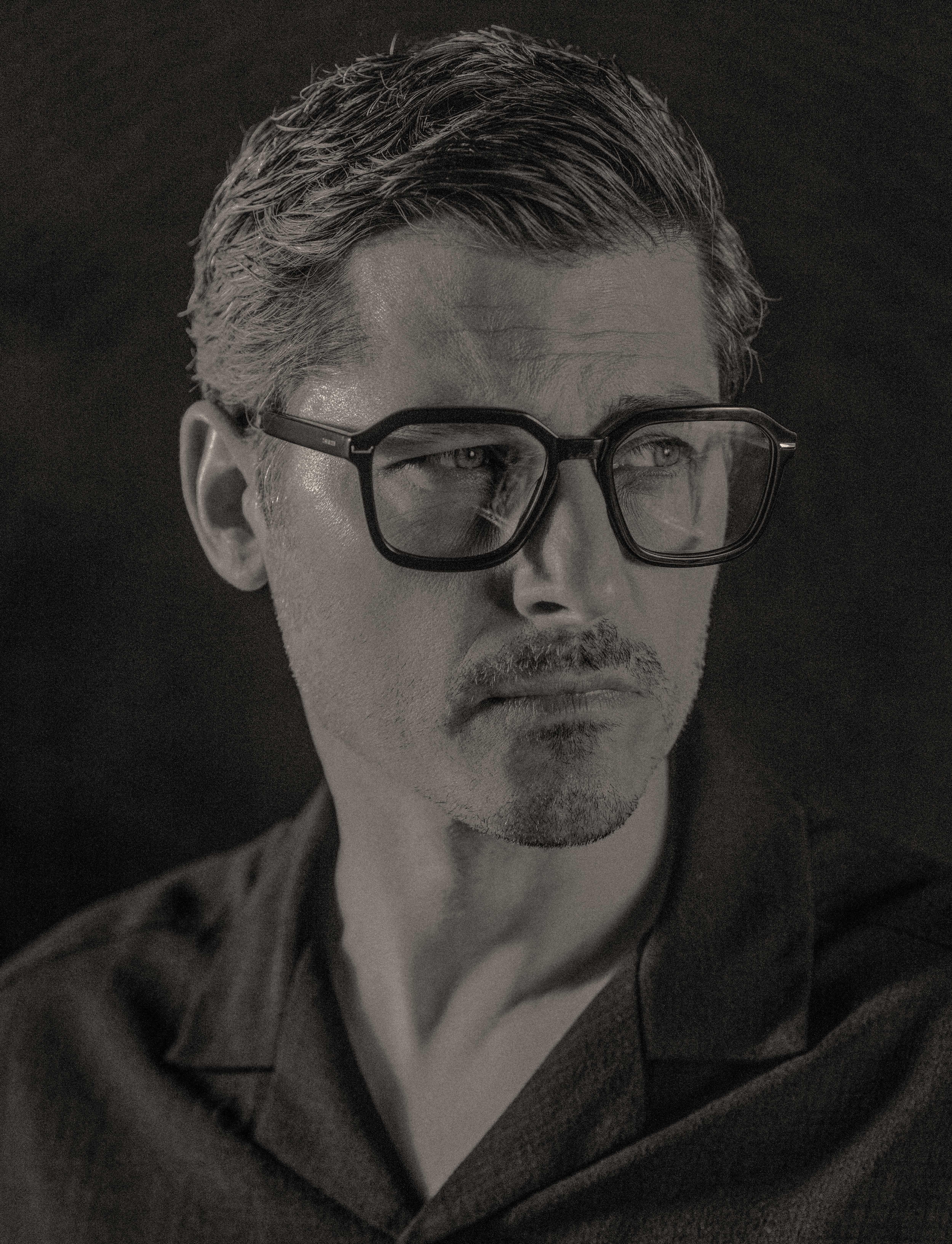 black and white portrait of a man with black optical glasses and black shirt in front of dark wall.