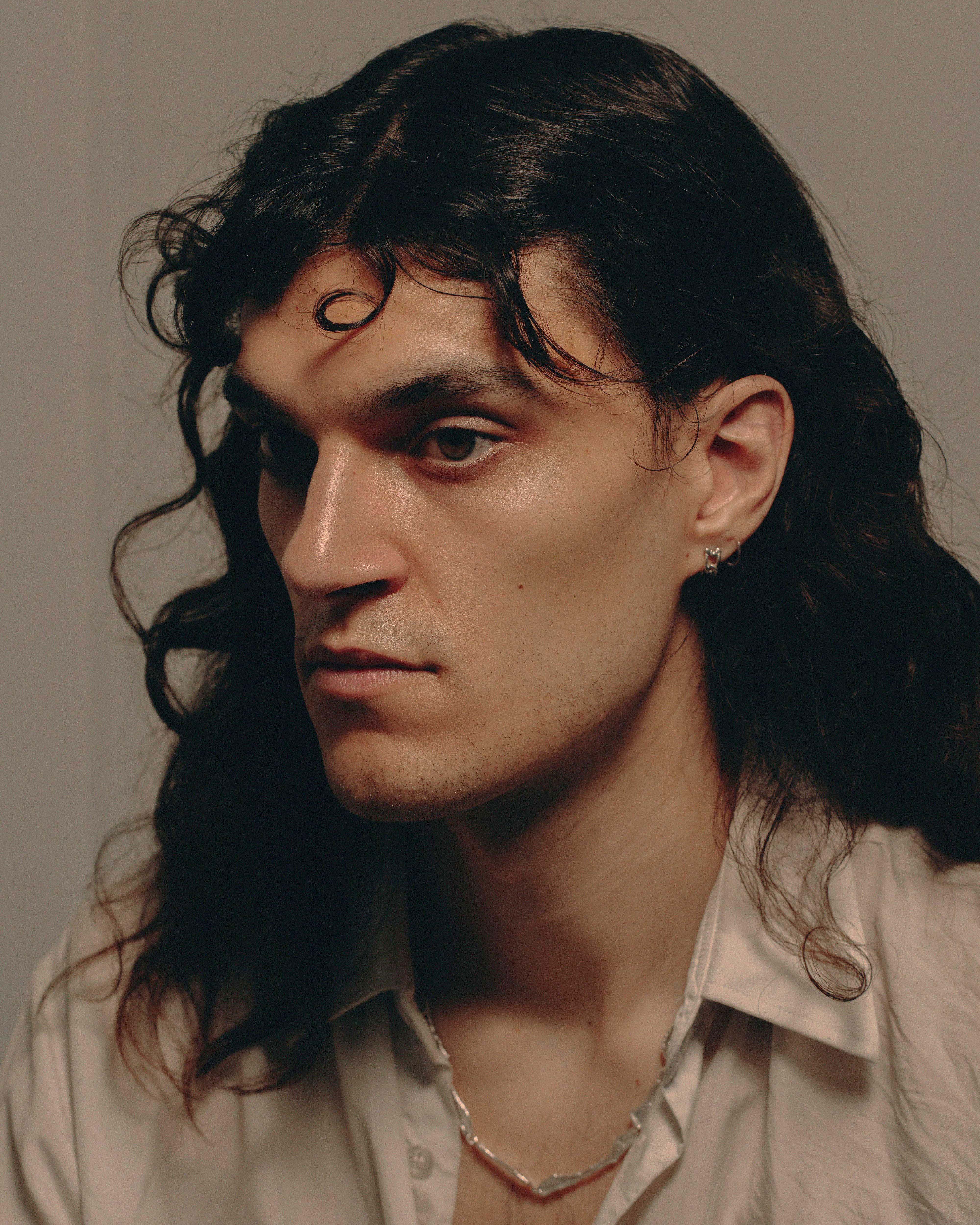 studio portrait of a young man with dark shoulder length hair in a light shirt with a silver chain around his neck.