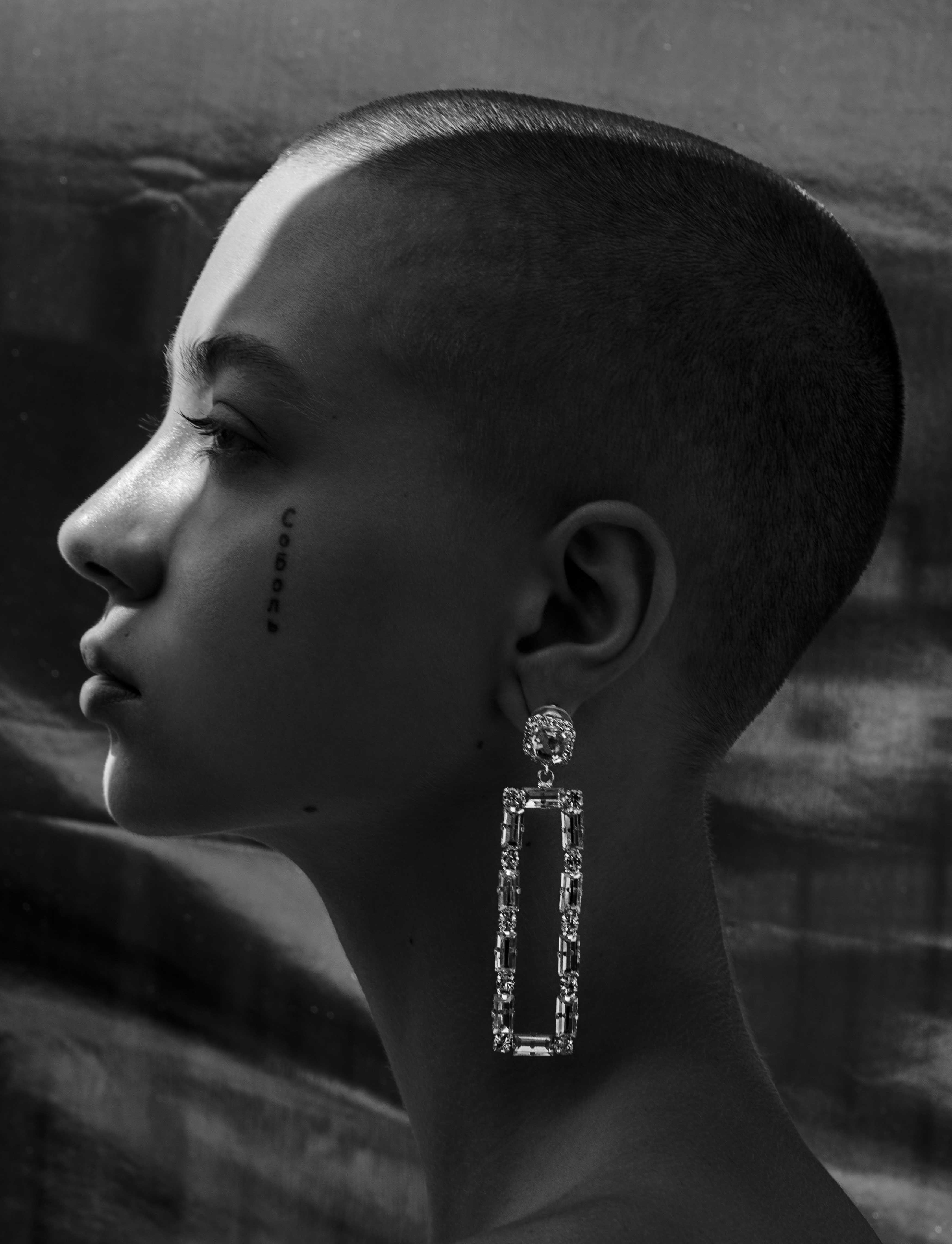 portrait of a woman with short shaved hair, a tattoo on her face and a long shiny earring. 