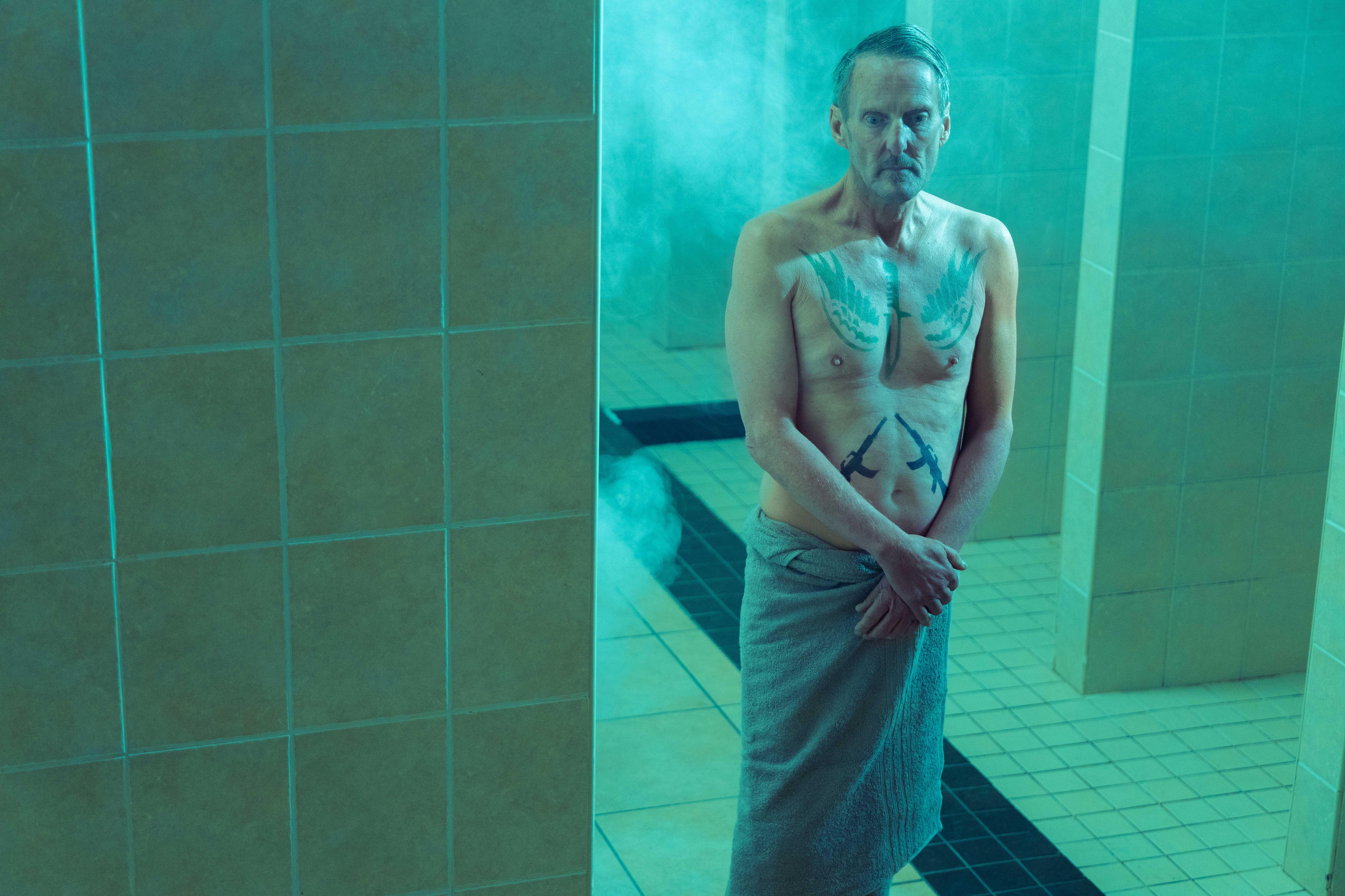 an older man stands in a washroom dressed only in a towel. he has tattoos on his chest.