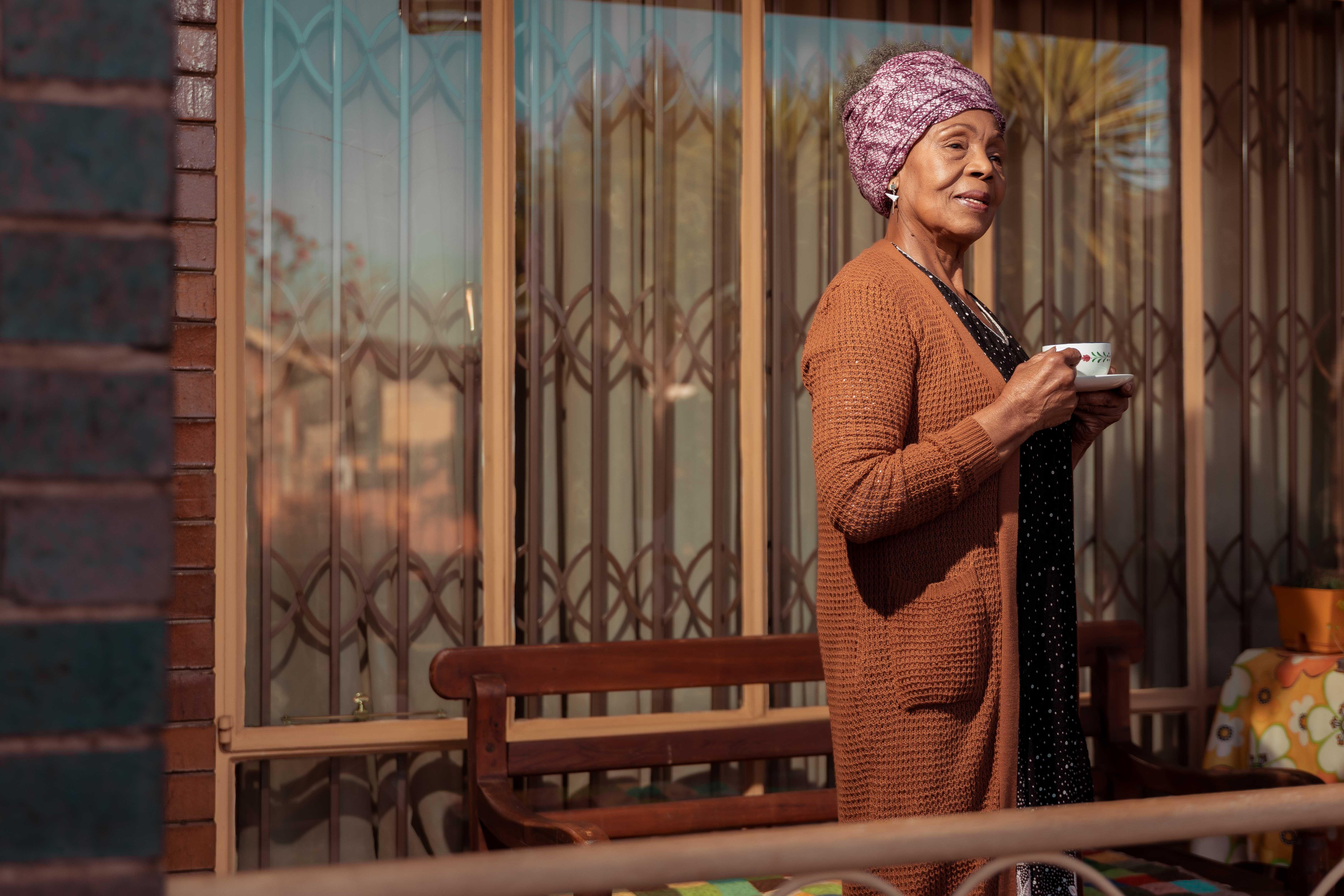 an african american elderly woman stands on the porch of her home with a cup of tea.