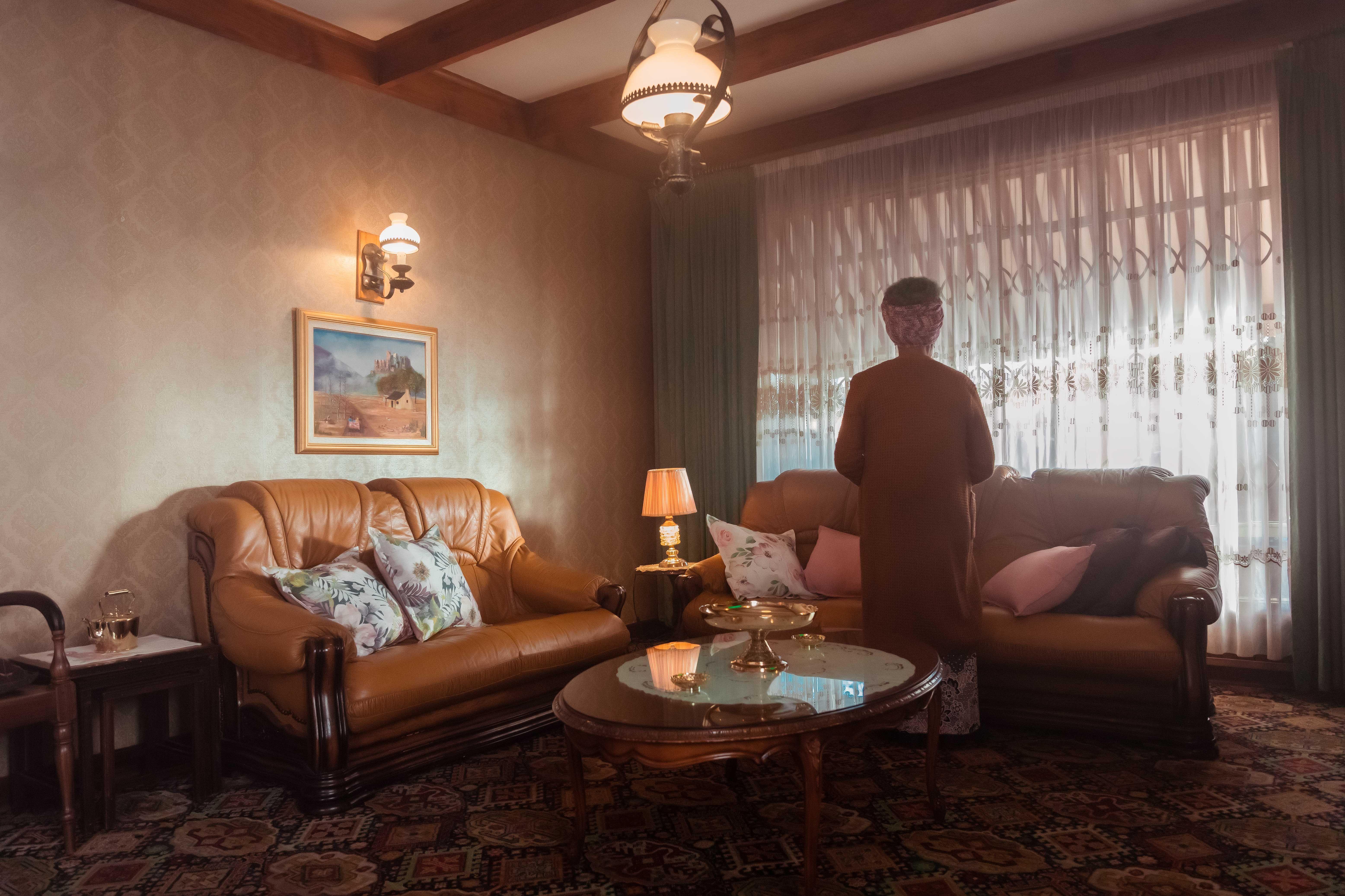 an elderly lady stands in her living room and looks out of the window.