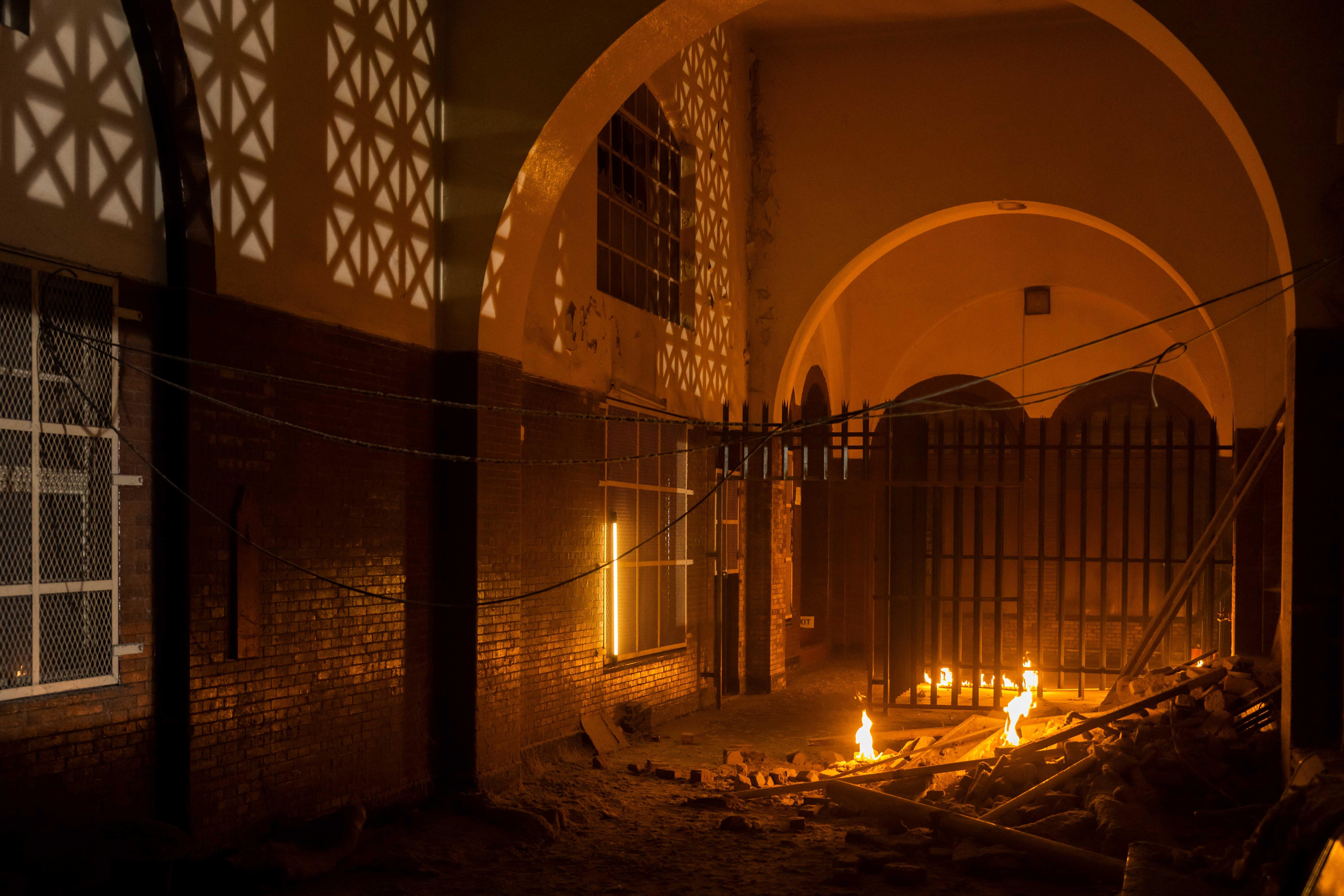 a dark hall with arches on the ceiling where debris and ashes lie and several small fires burn. 