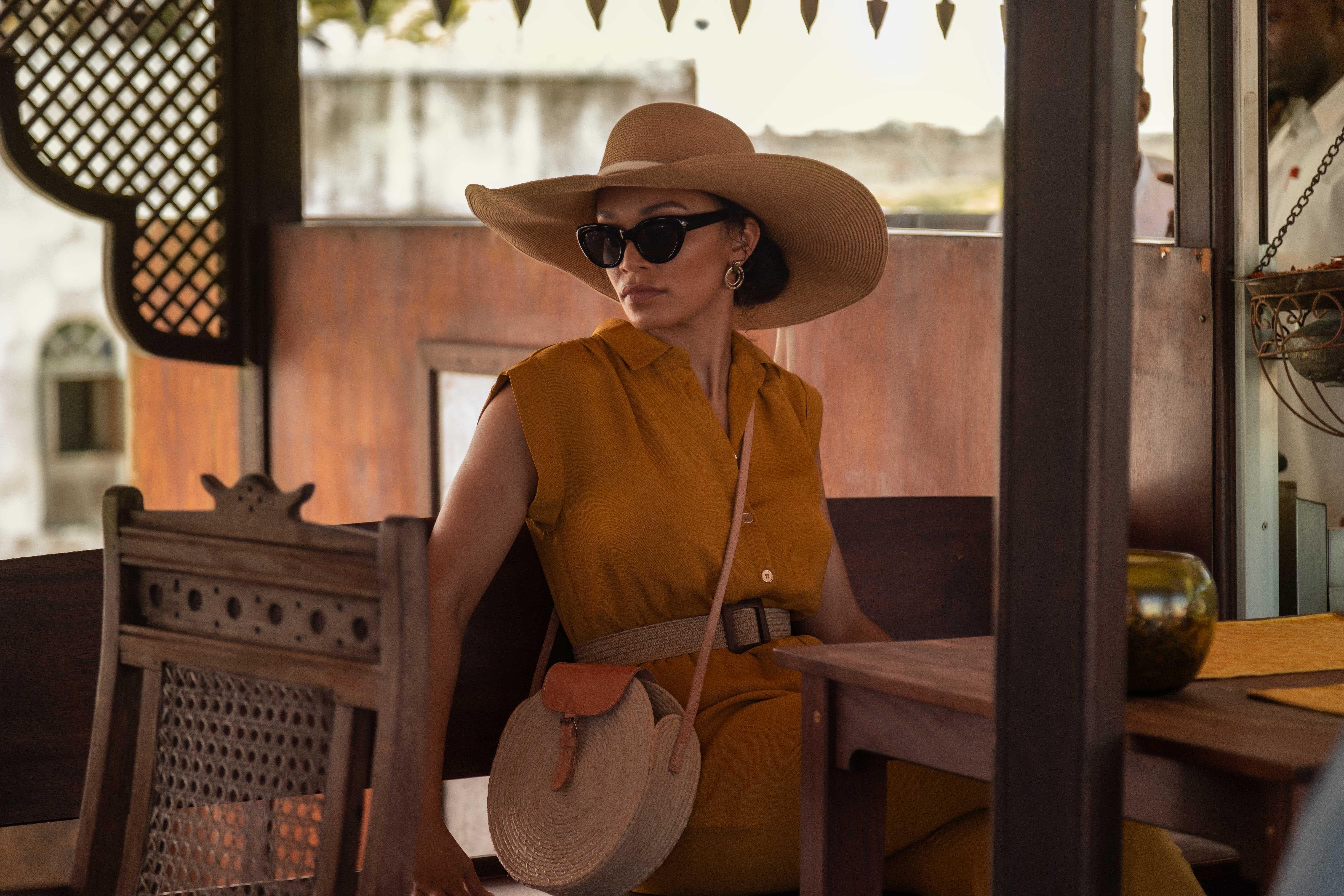 a woman with a hat and sunglasses sits at a table in a restaurant and watches the action.