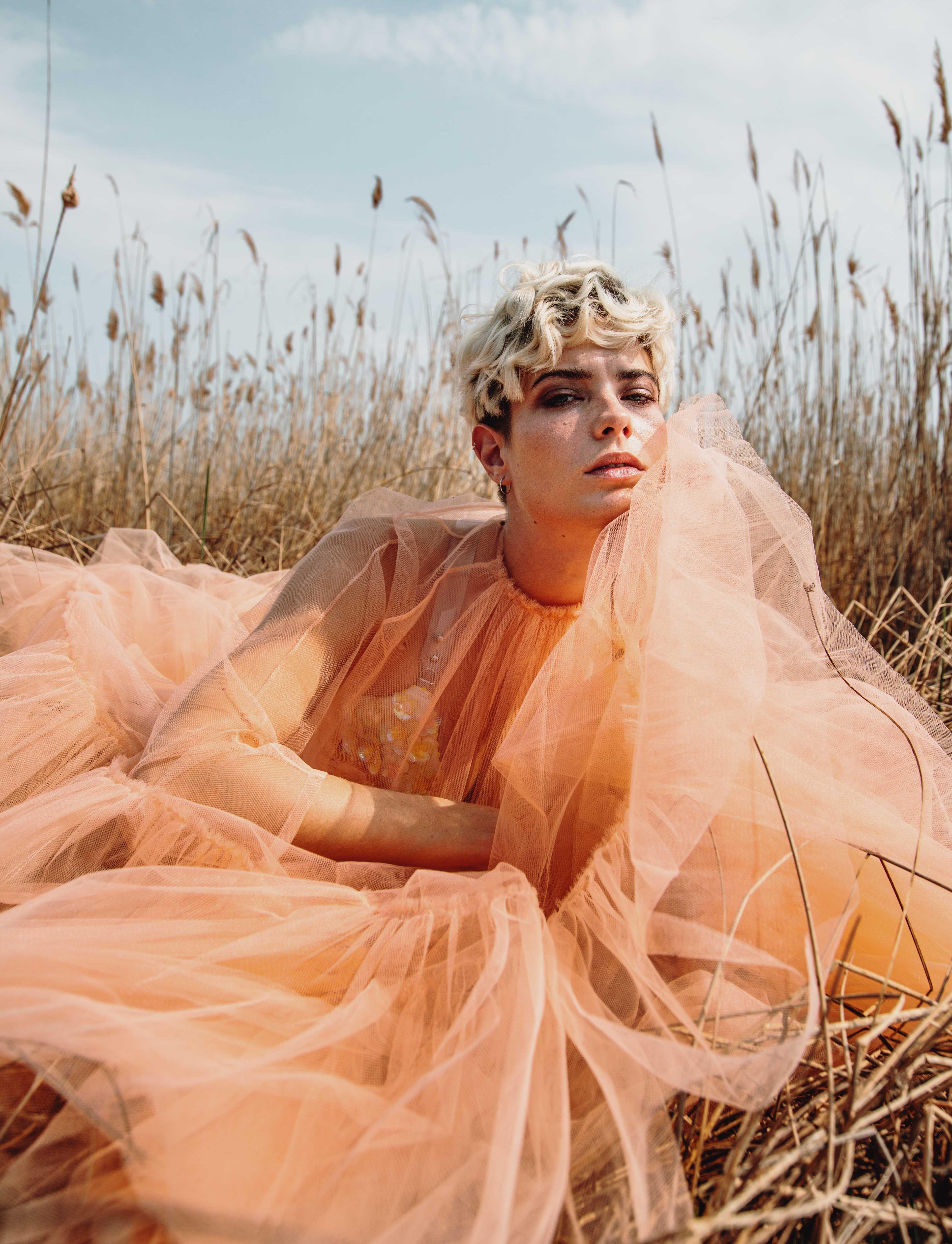 a young woman sits in a wheat field in a full-length tulle dress and looks bored into the camera.