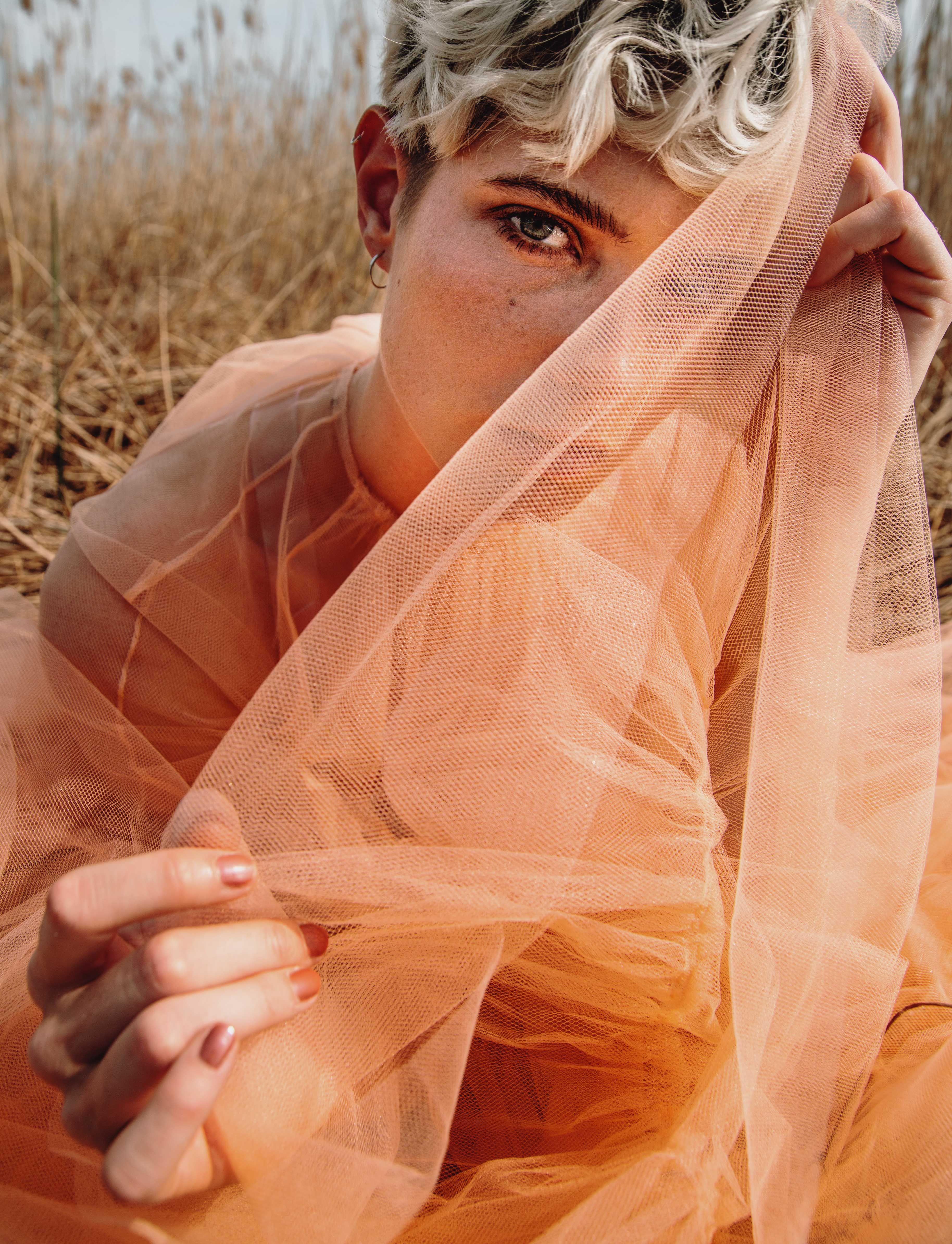 a young woman sits in a wheat field in a full-length tulle dress and plays with a piece of her dress and her look into the camera.